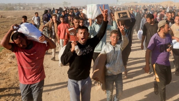 A group of people carrying boxes and holding up peace signs on sandy terrain.
