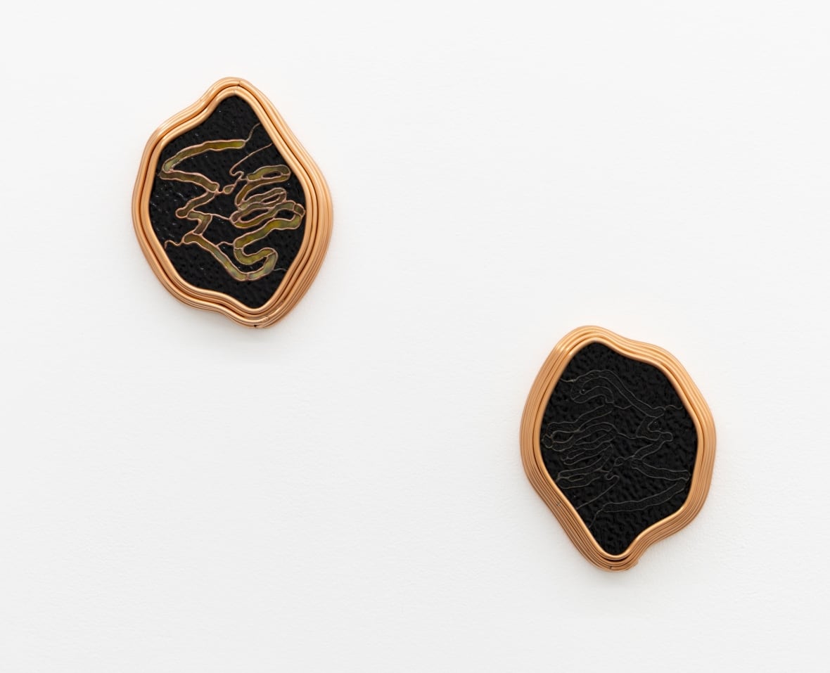 Two stained glass artworks hang on a white wall. One is black with serpentine cream-coloured markings. The other is black with just the outlines of markings visible.