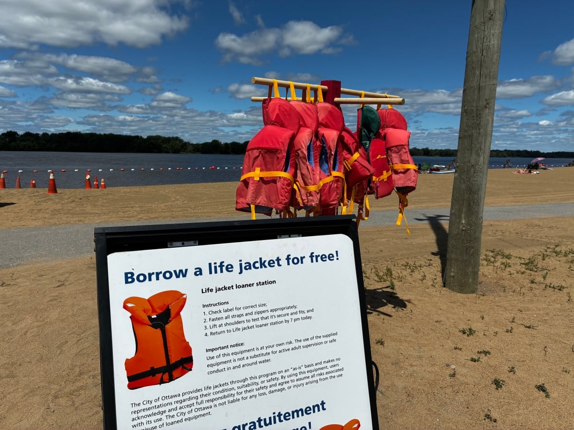 Red life jackets hang off of a wooden coat hangar on a beach.