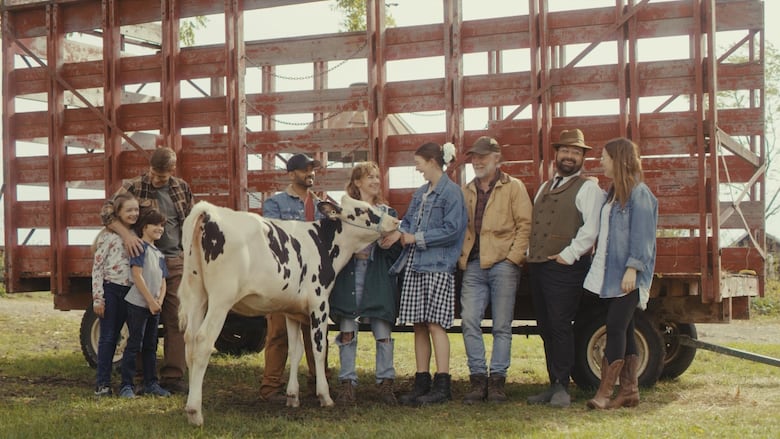 A diverse group of eight people, smiling, with a black and white calf in front of a large red farm trailer. They are in a grassy outdoor setting