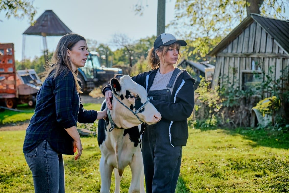 Two women, Allie Dunbar (left) and Lindsey Middleton (right), stand with a black and white cow in a grassy field on a farm set.