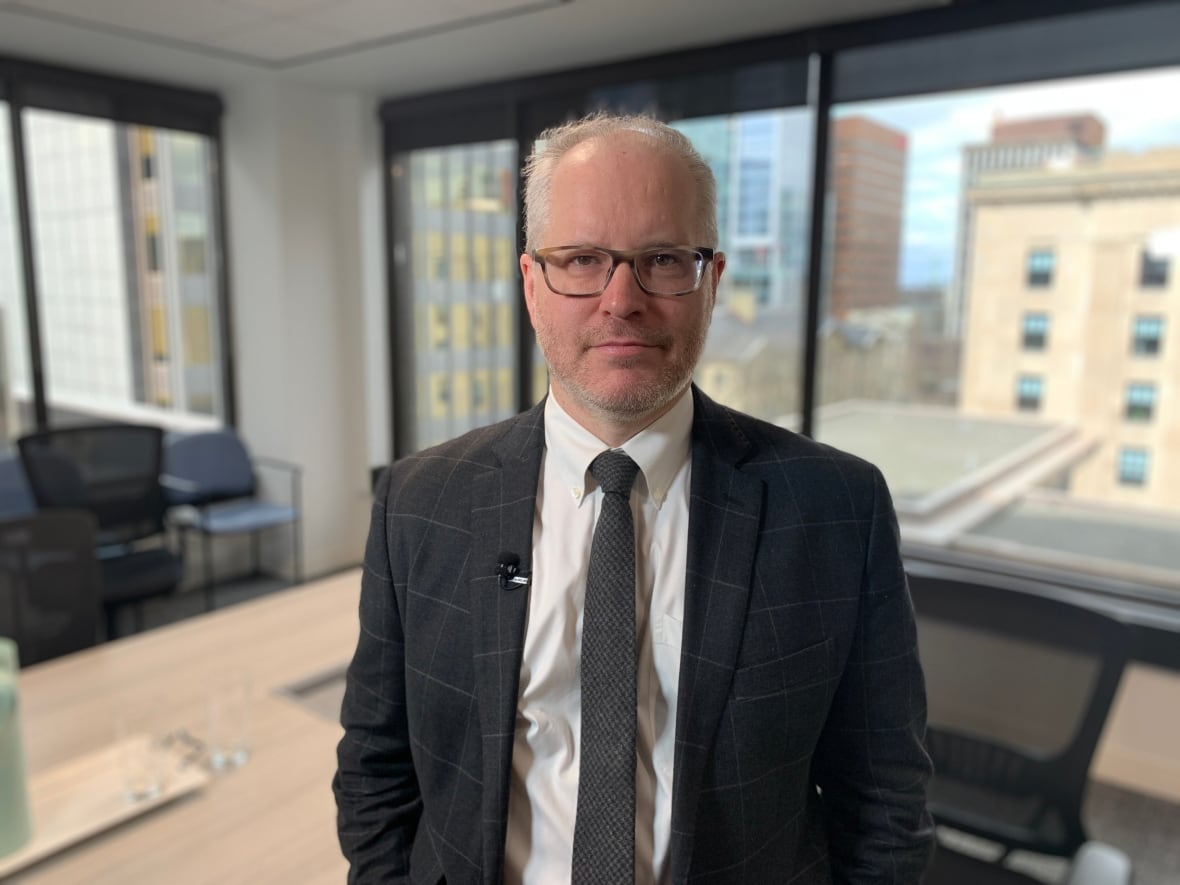 A portrait of a man wearing glasses and a suit, standing in a boardroom.