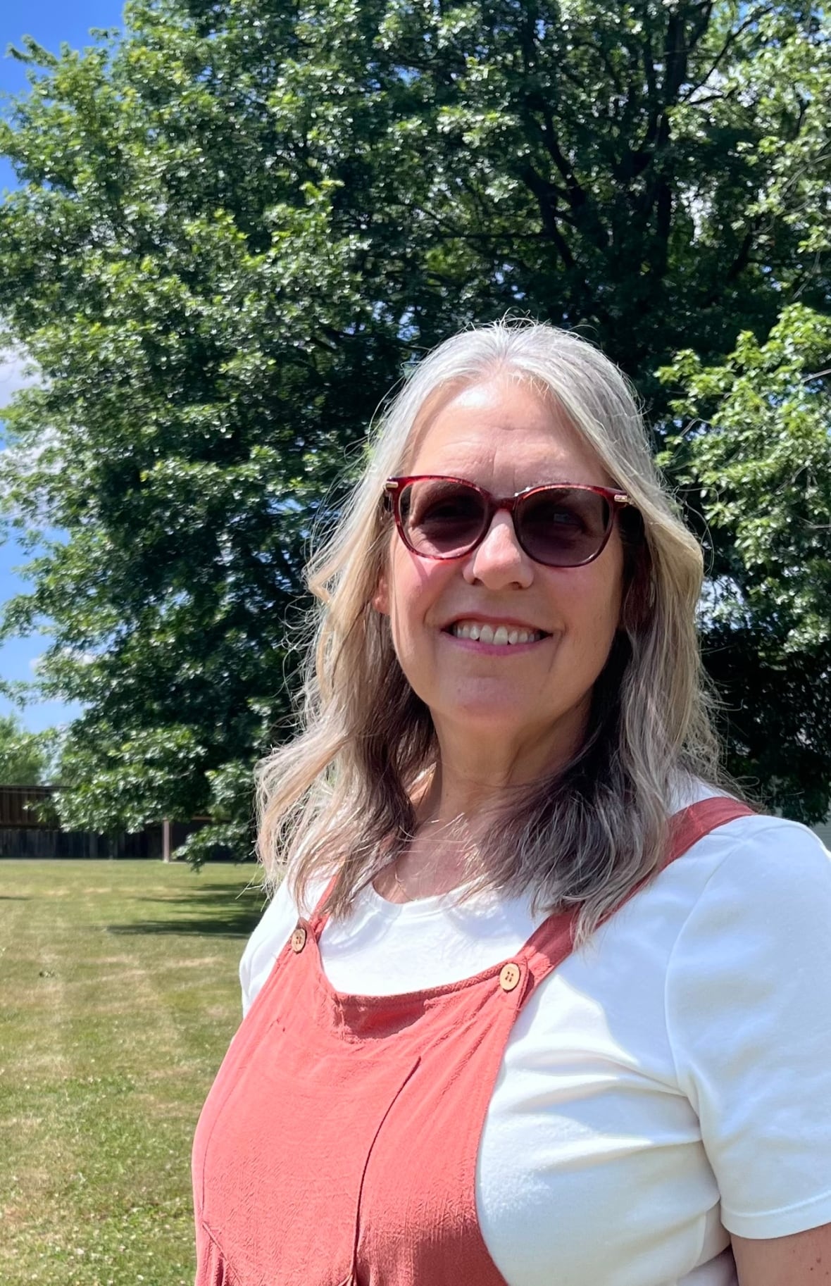 A smiling woman with light-colored hair and sunglasses, wearing a rust-colored top, standing outdoors in front of green trees.