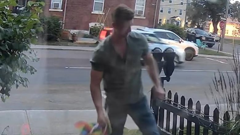 A man in a green shirt and blue pants stands on a path way, rainbow flags can be seen in his right hand.