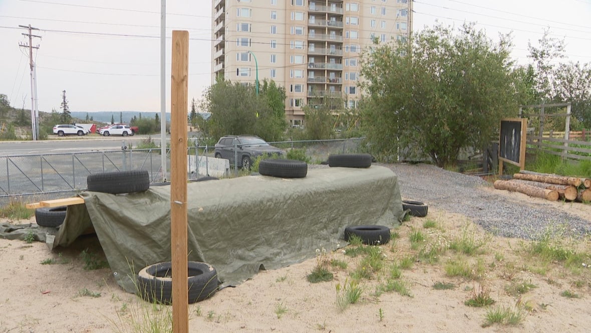 A schoolyard area under construction with logs, tires, and a structure covered by a tarp.