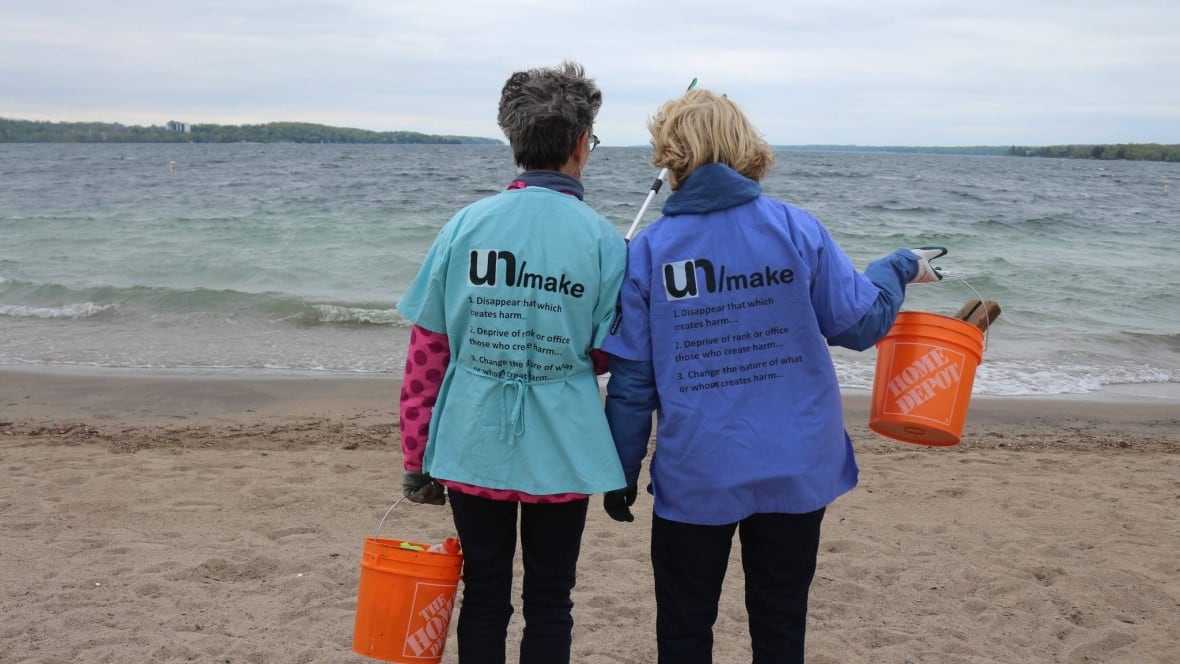 Two people wearing T-shirts that read "unMake" stand shoulder to shoulder on the sand facing the lake. They hold orange buckets.