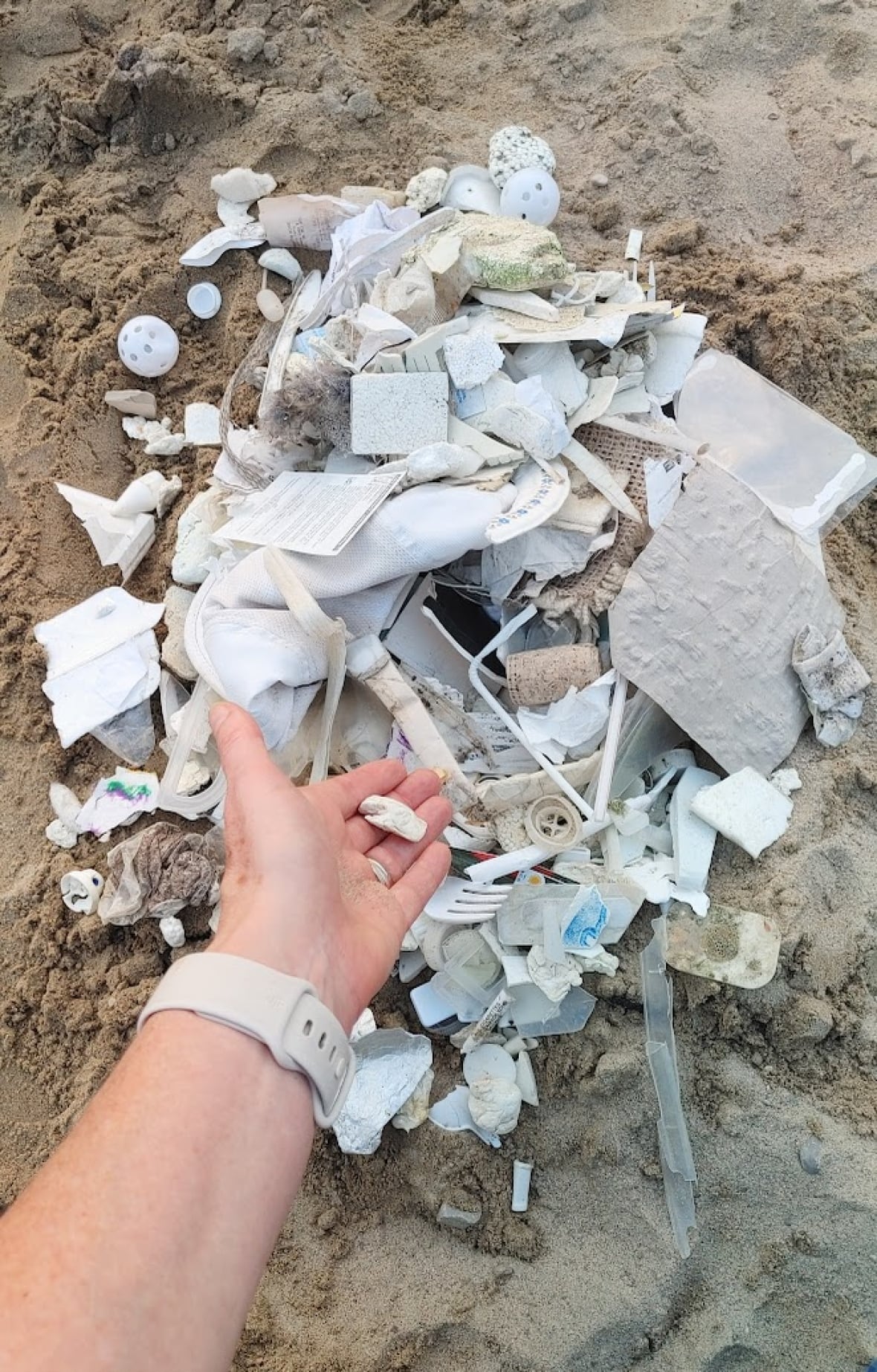 A hand gestures at a pile of white garbage forming a pile on the sand.