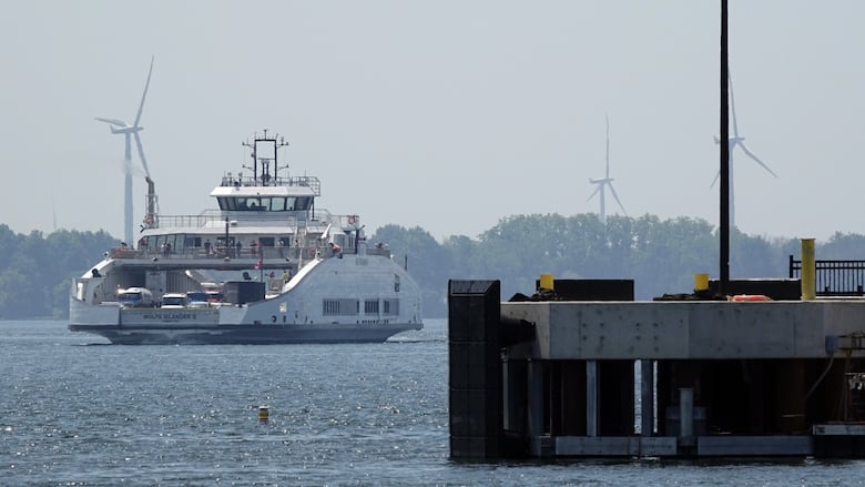 A large white boat carrying cars and people is shown rounding a metal and concrete dock. Wind turbines are turning in the background.