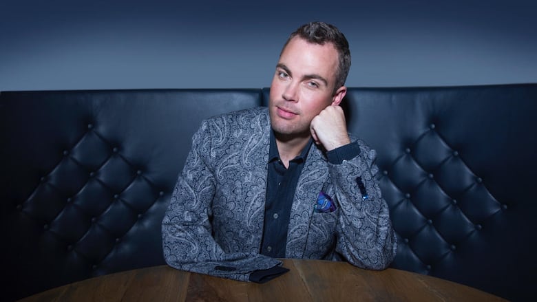 A man in a paisley blue suit jacket sits at booth smiling wryly with his face resting on his left hand. His jacket's empty right sleeve is laid flat on the table in front of him. 