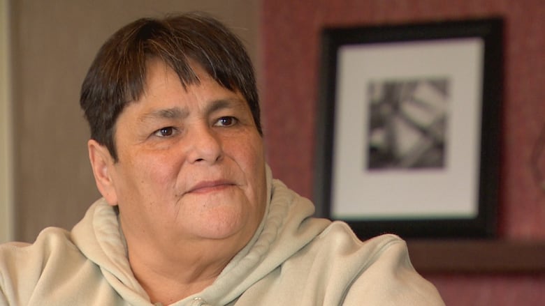 A light-skinned woman, with short brown hair, is wearing a stone-coloured hoodie. She is sitting in room with red wallpaper.