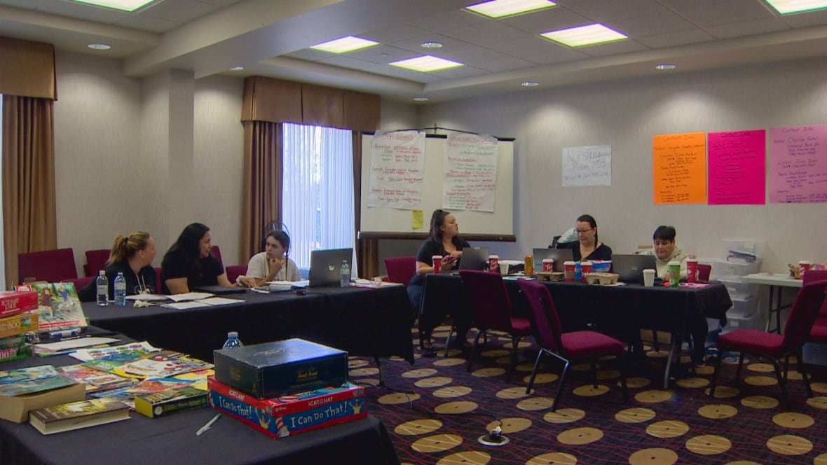 Six women sit at two folding tables in a carpeted boardroom. Bristol board and large sheets of paper are posted on a whiteboard and a wall.