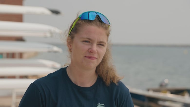 A woman with her blonde hair in a ponytail squints at the camera. She is wearing a T-shirt, there are sunglasses on her head, and there are several rowing boats behind her.