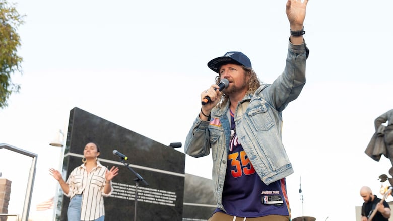 A casually dressed man sings into a microphone with one arm raised in the air.