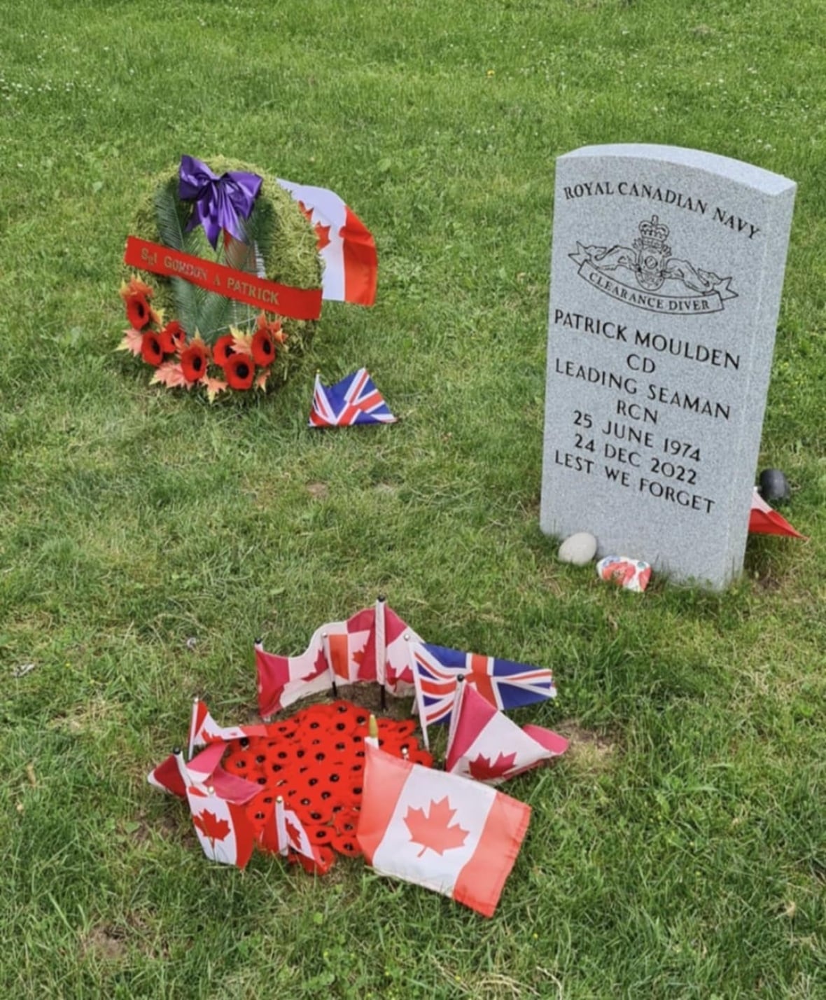 A military headstone with Patrick Moulden's name on it, next to it is a green wreath with flowers on it, and the name Gordon Patrick.