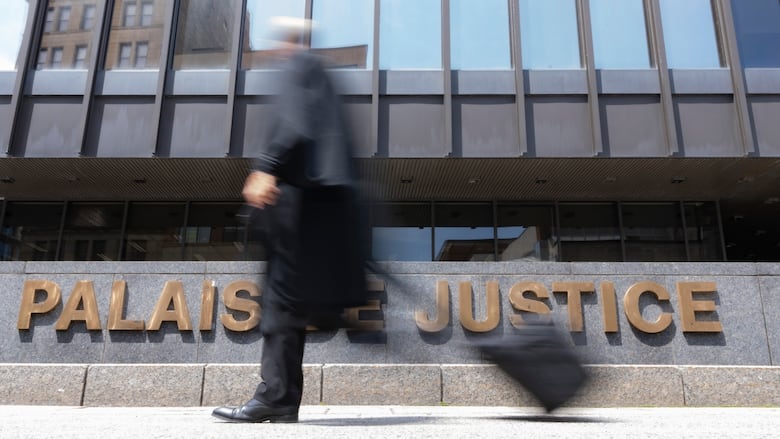 A man wearing black robes and carrying a suitcase is out of focus in front of a large building.