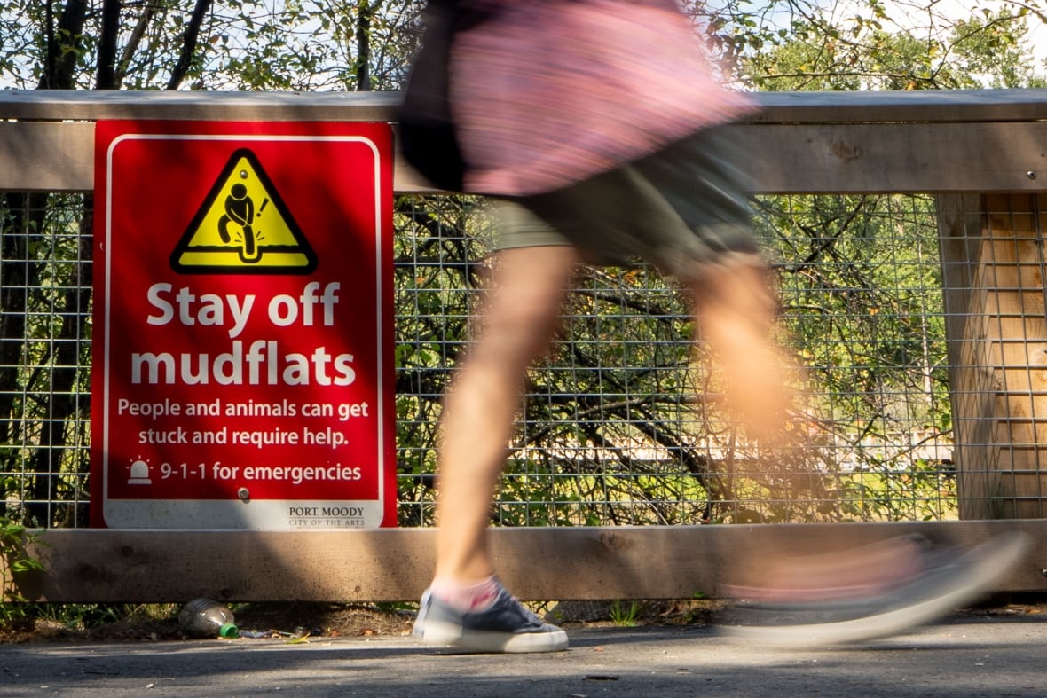 A person walks past a sign that reads 'Stay off mudflats. People and animals can get stuck and require help. 911 for emergencies'.