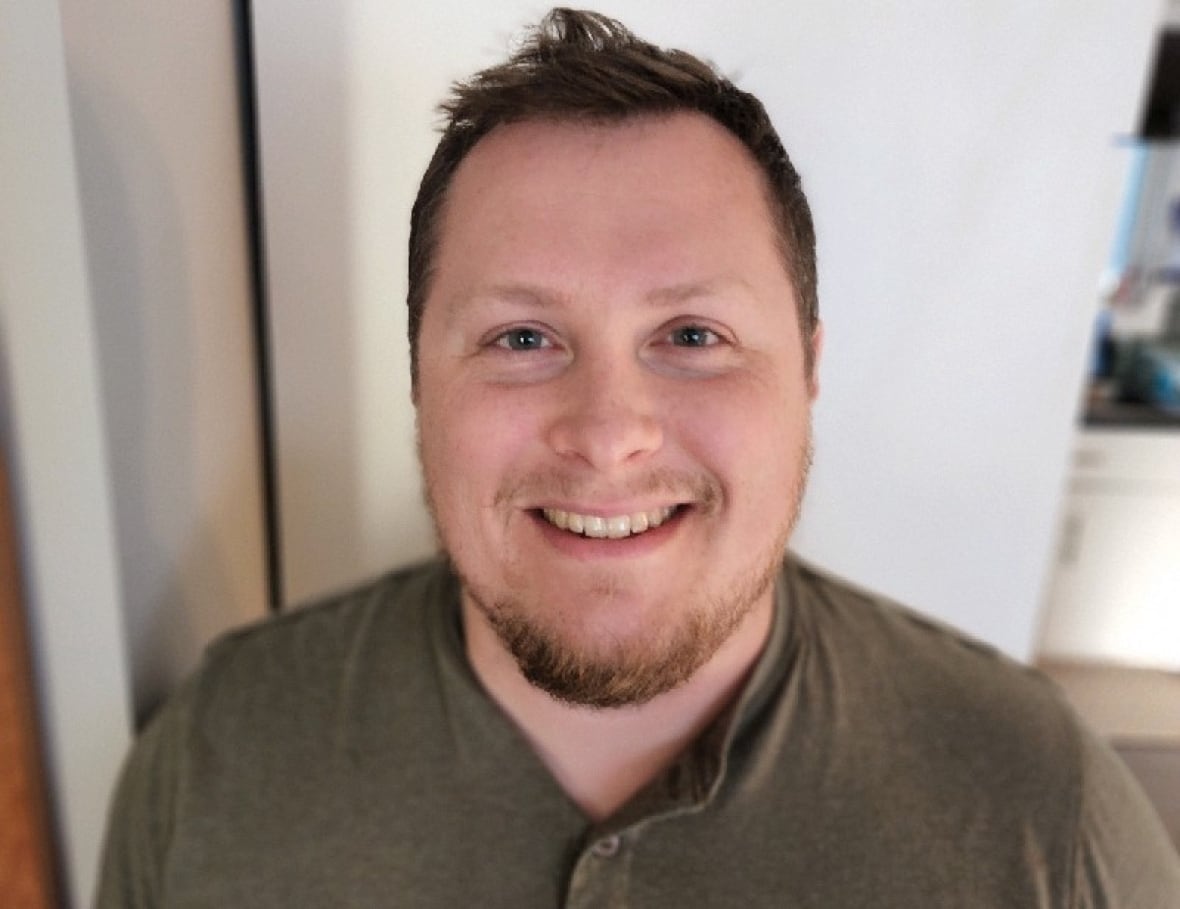 A man with short hair and a beard, wearing a green henley shirt, smiling in a well-lit indoor setting