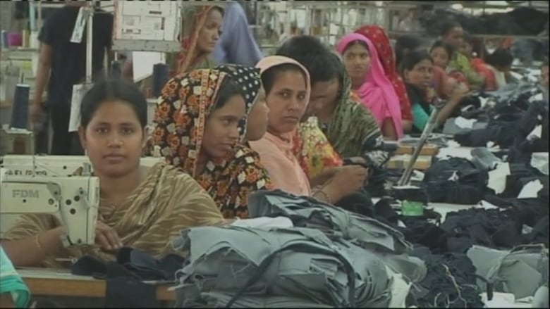 Several women wearing sarees sit in a row behind each other, with a sewing machine in front of them and piles of fabric next to them.