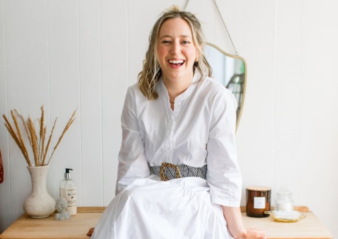 A woman in a white dress sits barefoot on a wooden dresser, smiling, with a mirror and decorative items in the background
