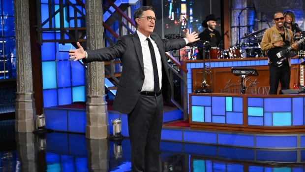 A man in a black suit, white shirt, black tie and black glasses stands with his arms stretched out on a TV set with a band in the background. 