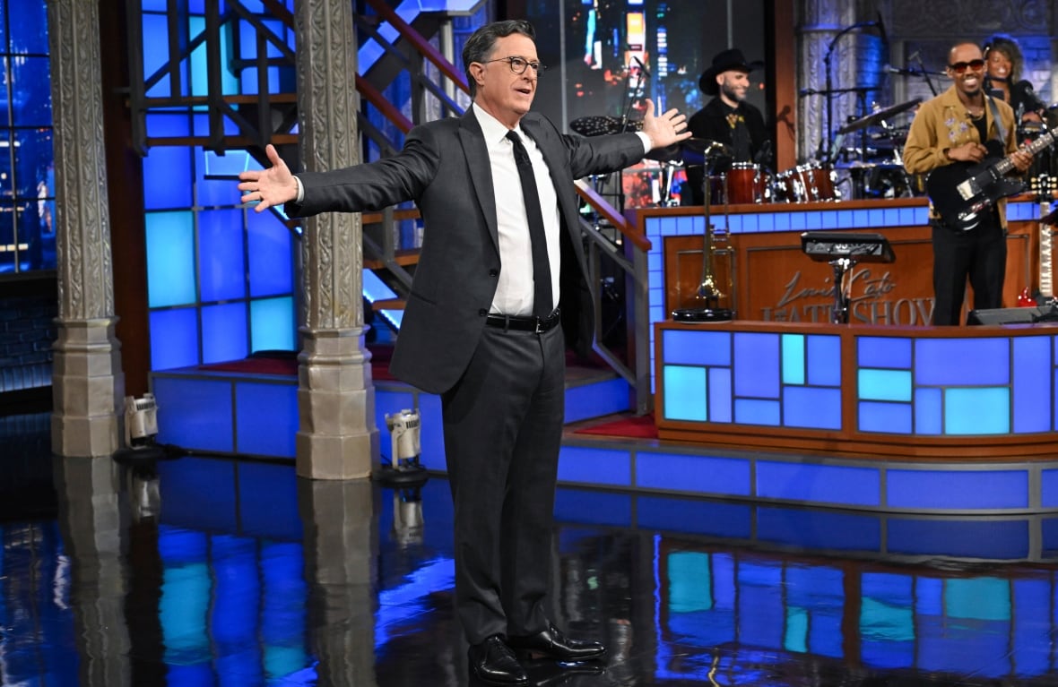 A man wearing a black suit, a white shirt, a black tie and dark glasses standing with arms stretched on TV in the band is set in the band. 