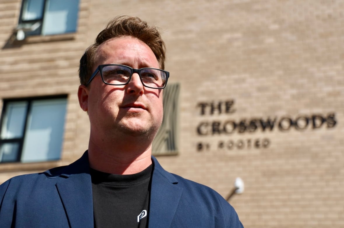 A portrait of a man wearing glasses in the sun with a brick building in the background.