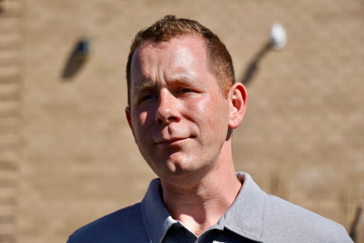 A portrait of a man in the sun with a brick building in the background.