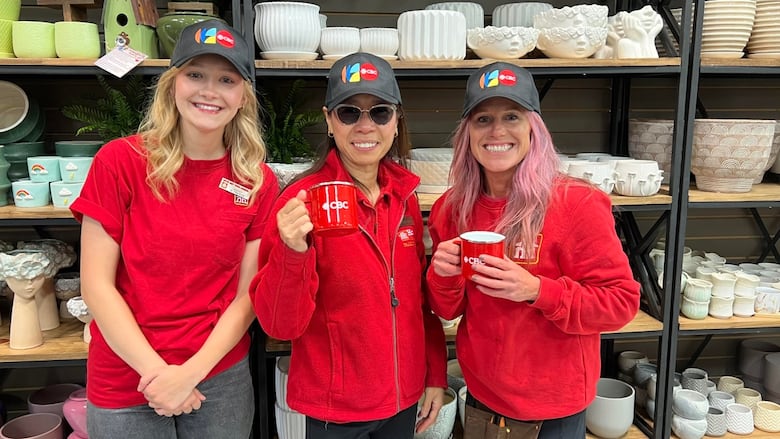 3 women in red Home Hardware shirts smiling at the camera. They are holding red CBC mugs and wearing black CBC ball caps.