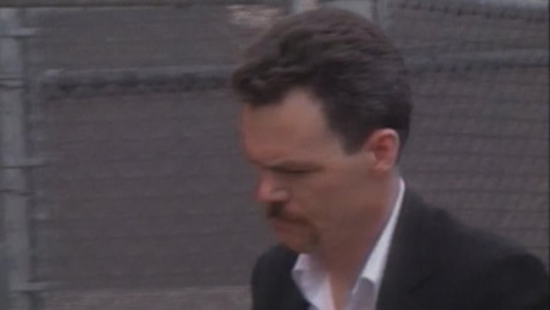 A man with brown hair and facial hair walks against a fence in a suit.