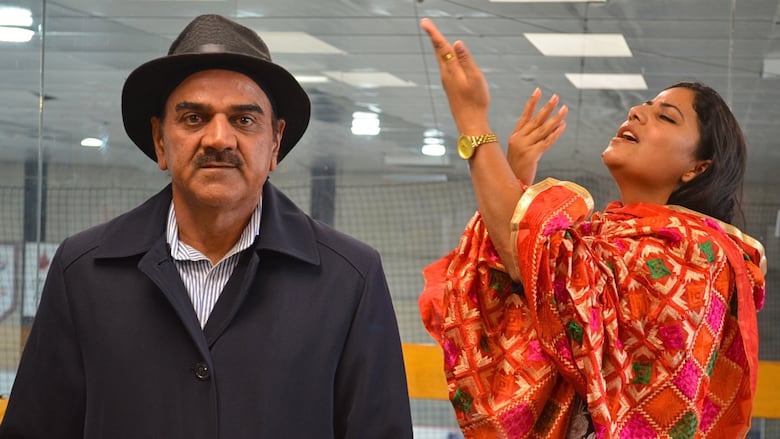 A man in a hat and coat stand looking directly at the camera. A woman wrapped in a vibrant red saree sings to him.