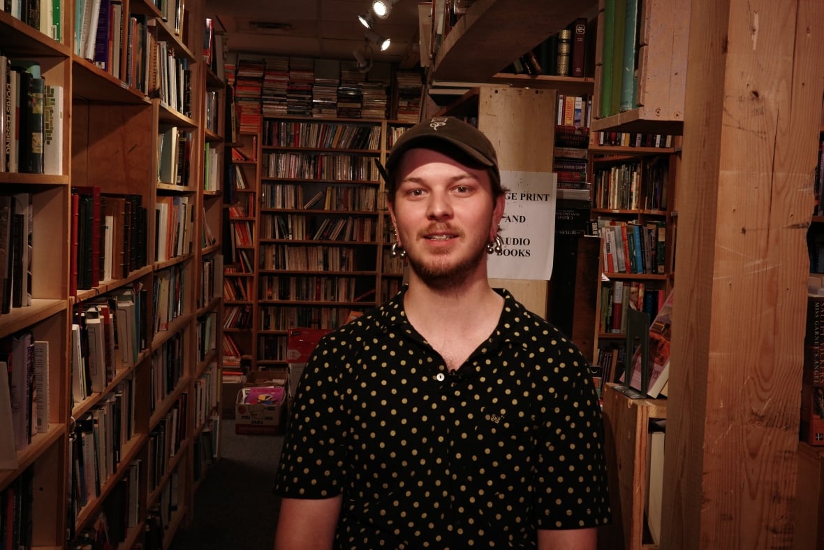 A man wearing a black polka dot shirt and a baseball cap poses for a photo. He is standing in front of several bookshelves. 