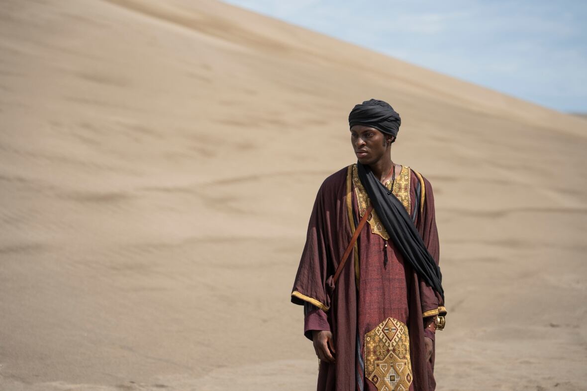 A young man wearing a robe and head scarf walks among sand dunes.