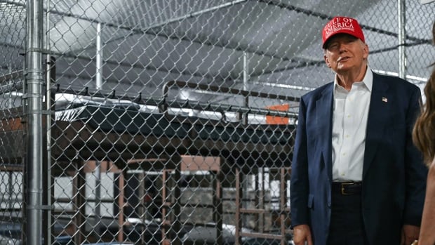 Donald Trump stands smiling in a suit and a red MAGA hat in front of empty prison cells with bunk beds, each separated by chicken wire 