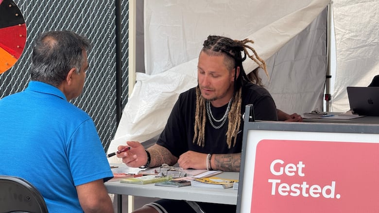 Two people sit across from each other at a table under a tent in a parking lot.