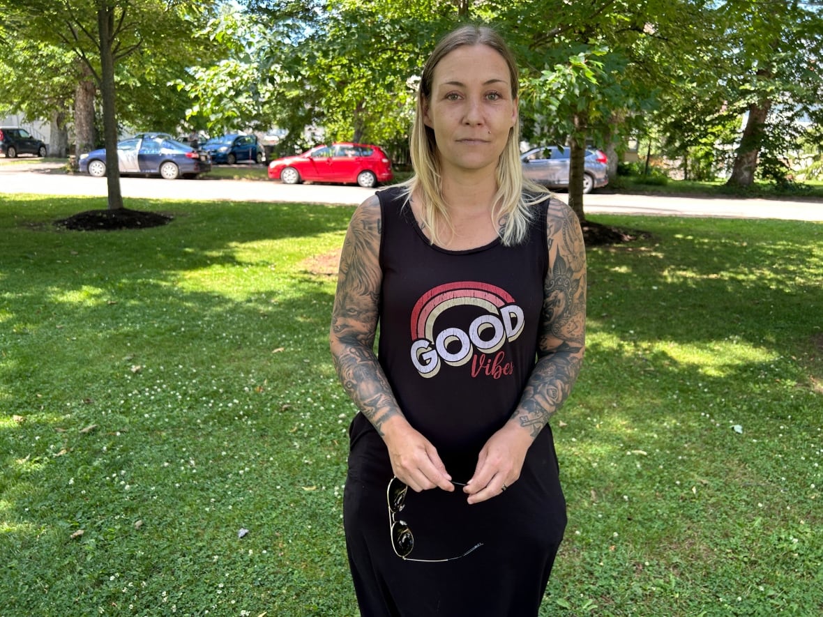A woman with blonde hair and a black tank top stands under two trees.