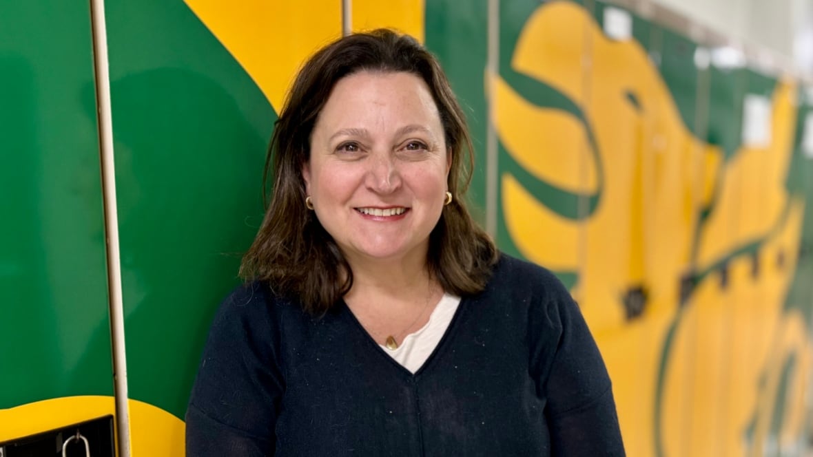 Woman wearing navy blue cardigan stands smiling next to a gold and yellow wall of painted lockers.
