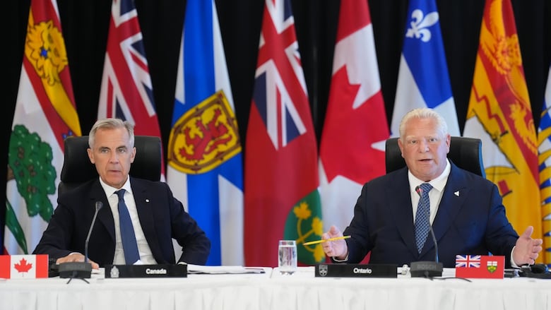 Two men sit at a table with flags behind them.