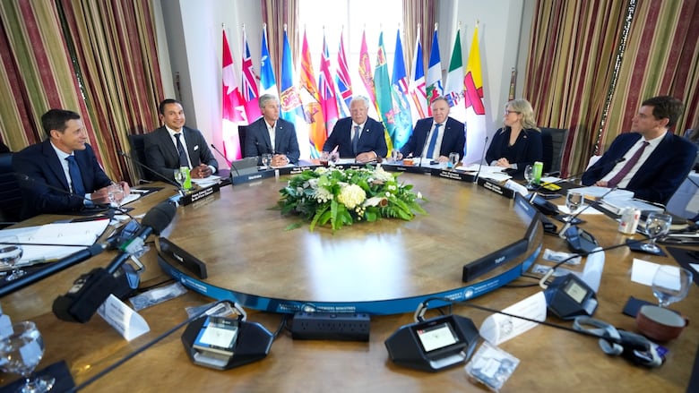 Group of people sit around round table wearing suits, with flags behind them