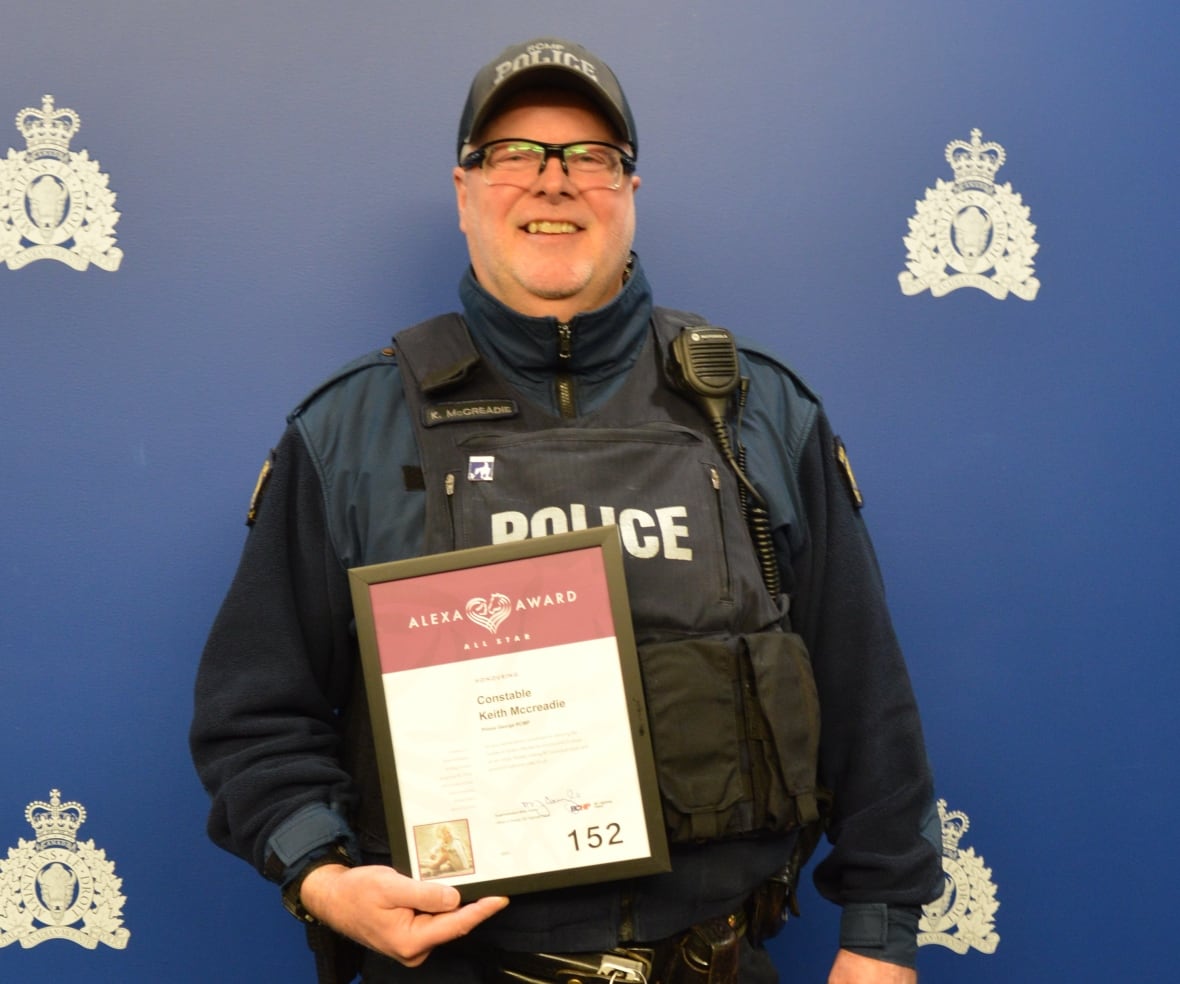 A smiling police officer wearing a vest that says, Police, across it, holds a plaque.