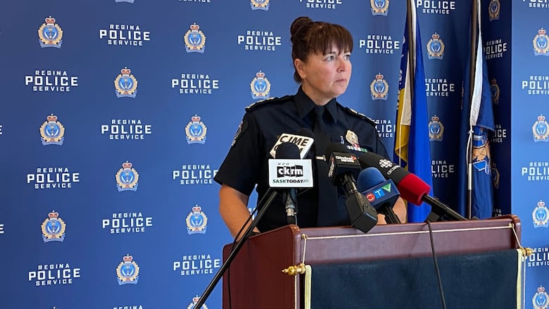 A female police officer in uniform speaks before a Regina Police Services podium and backdrop.  