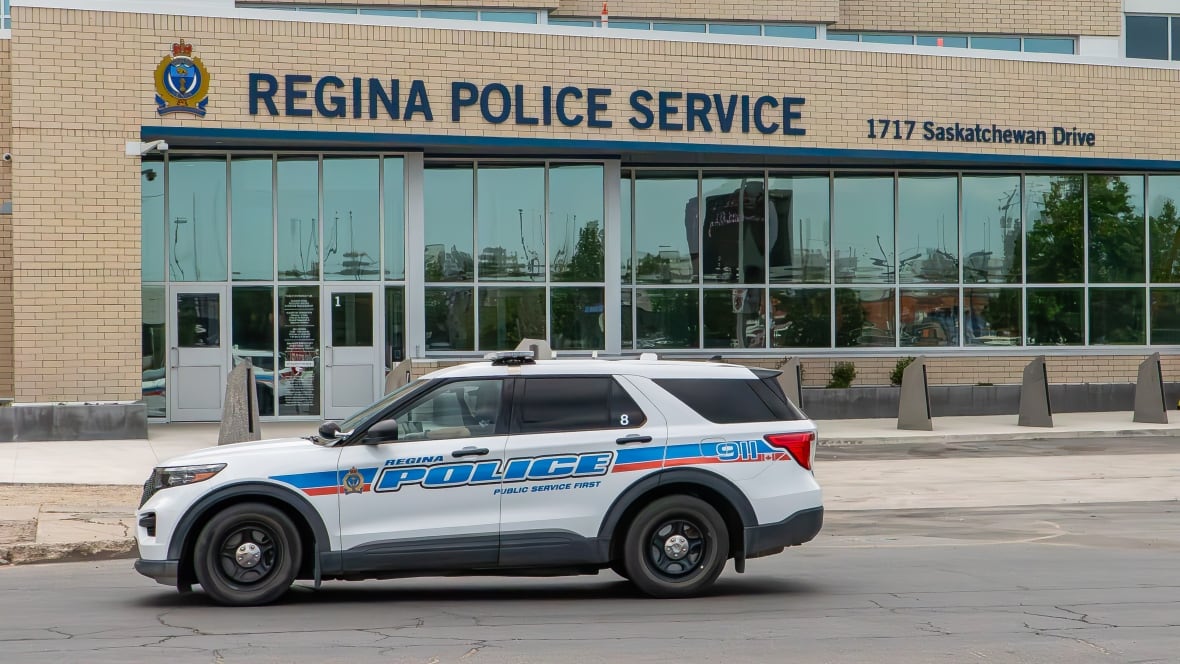 A white and blue police cruiser is parked in front of a tan brick building that says Regina Police Services.