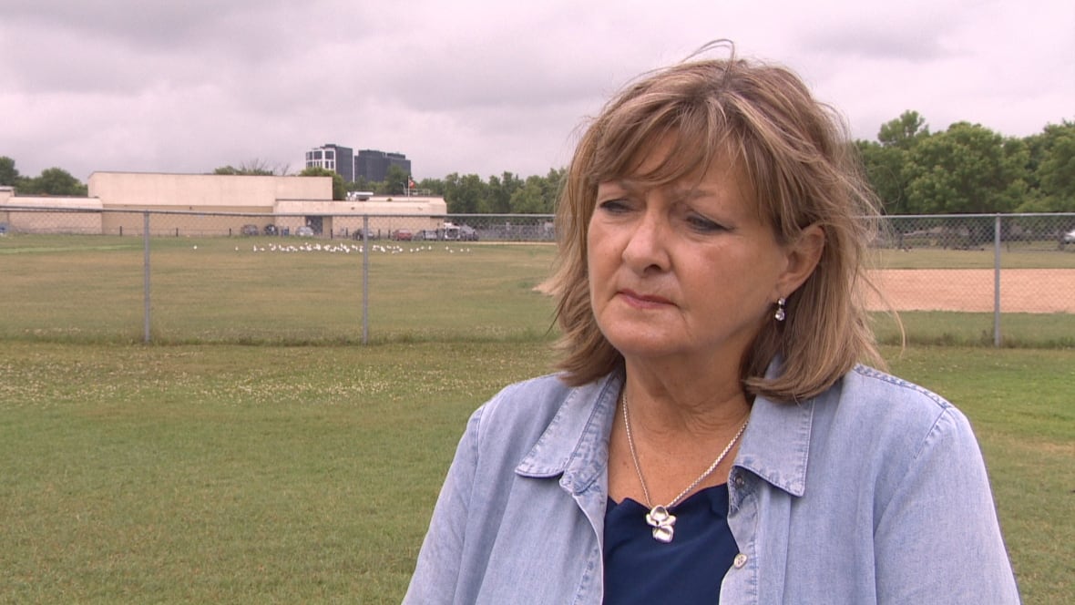 Woman with shoulder-length hair wears a blue button up shirt and stands in a field with short-cut grass.