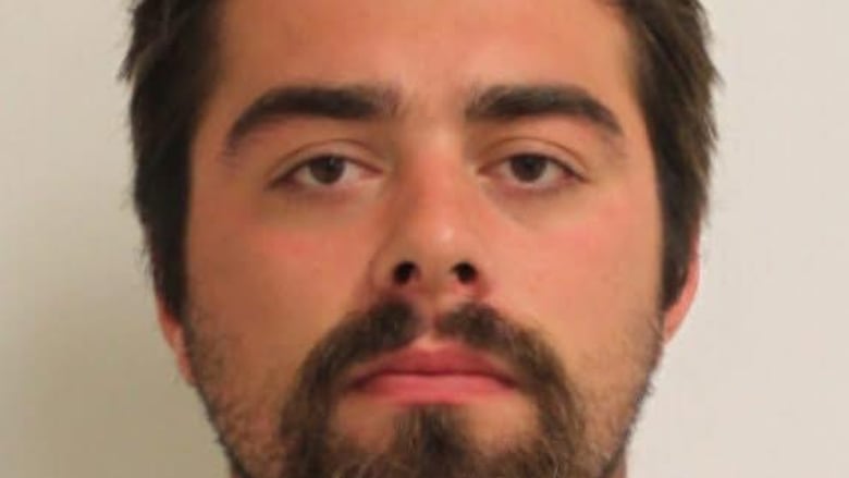 A headshot of Mitchell Lannigan, a man with a beard and dark hair, looking directly at the camera. He is wearing a grey and white hooded shirt