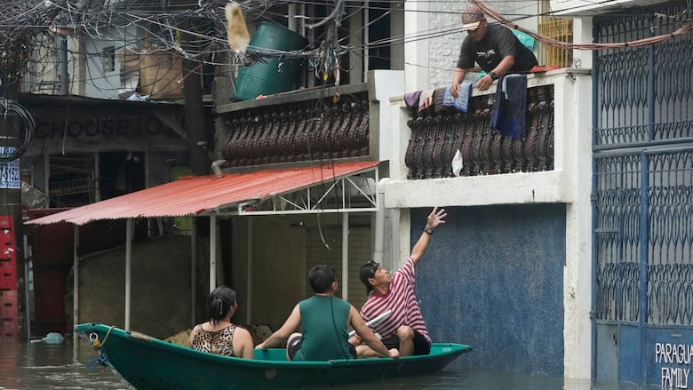 People sit in a green boat while a man on a balcony throws an item to one of them while floating in water on a street.