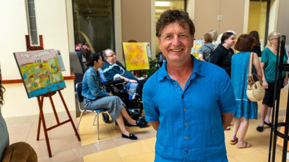 Christopher Griffin standing at the Hallways of Hope art exhibit, with vibrant patient-created paintings and attendees visible behind him.