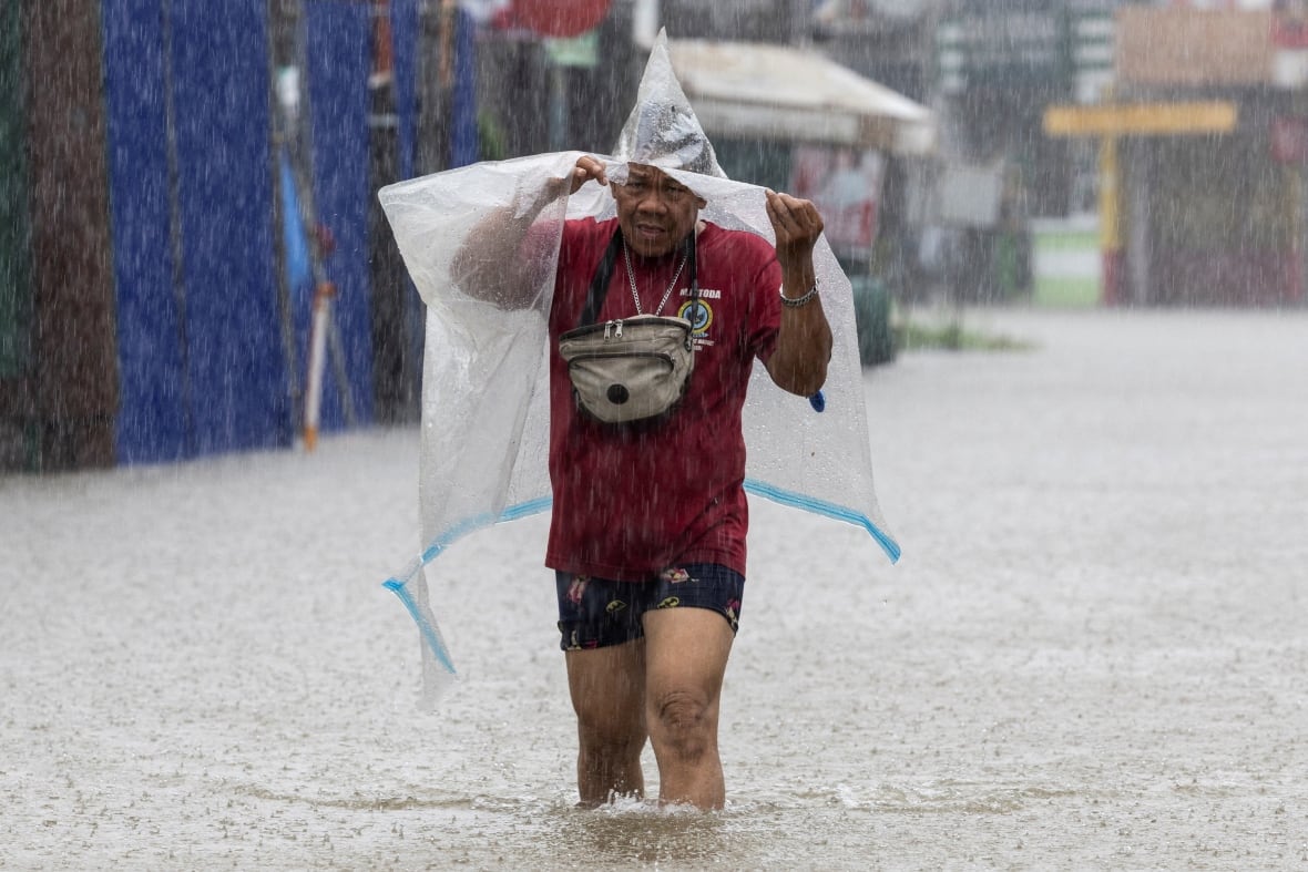 A man in a red shirt hgods a clear plastic tarp over his head as he walks down a street flooded with water up to his knees.