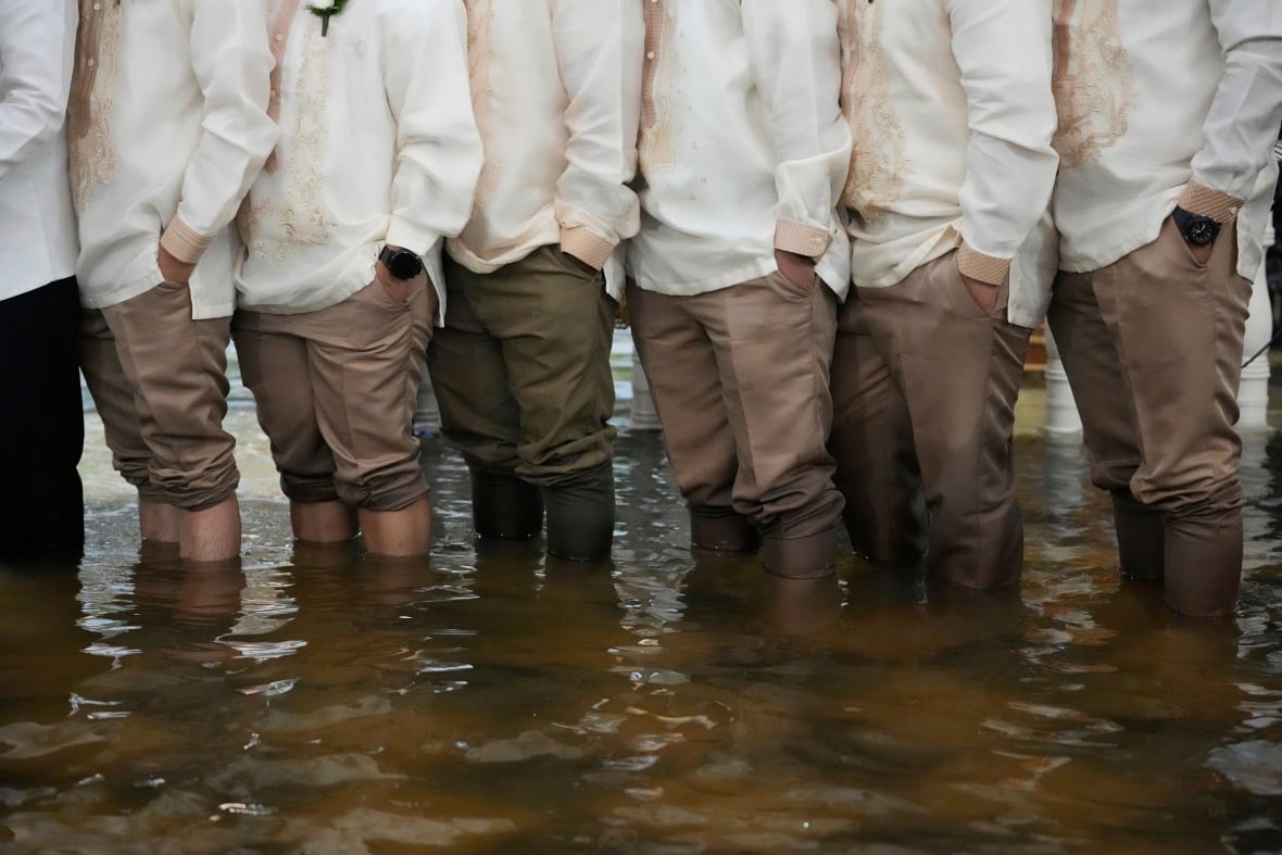 Men wearing brown pants rolled up past their ankles stand in murky brown water