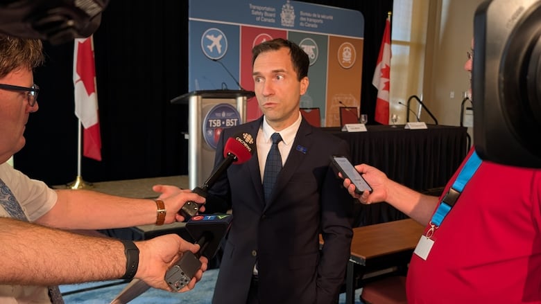 A man in a suit is being interviewed by reporters with microphones and a smartphone at a press conference