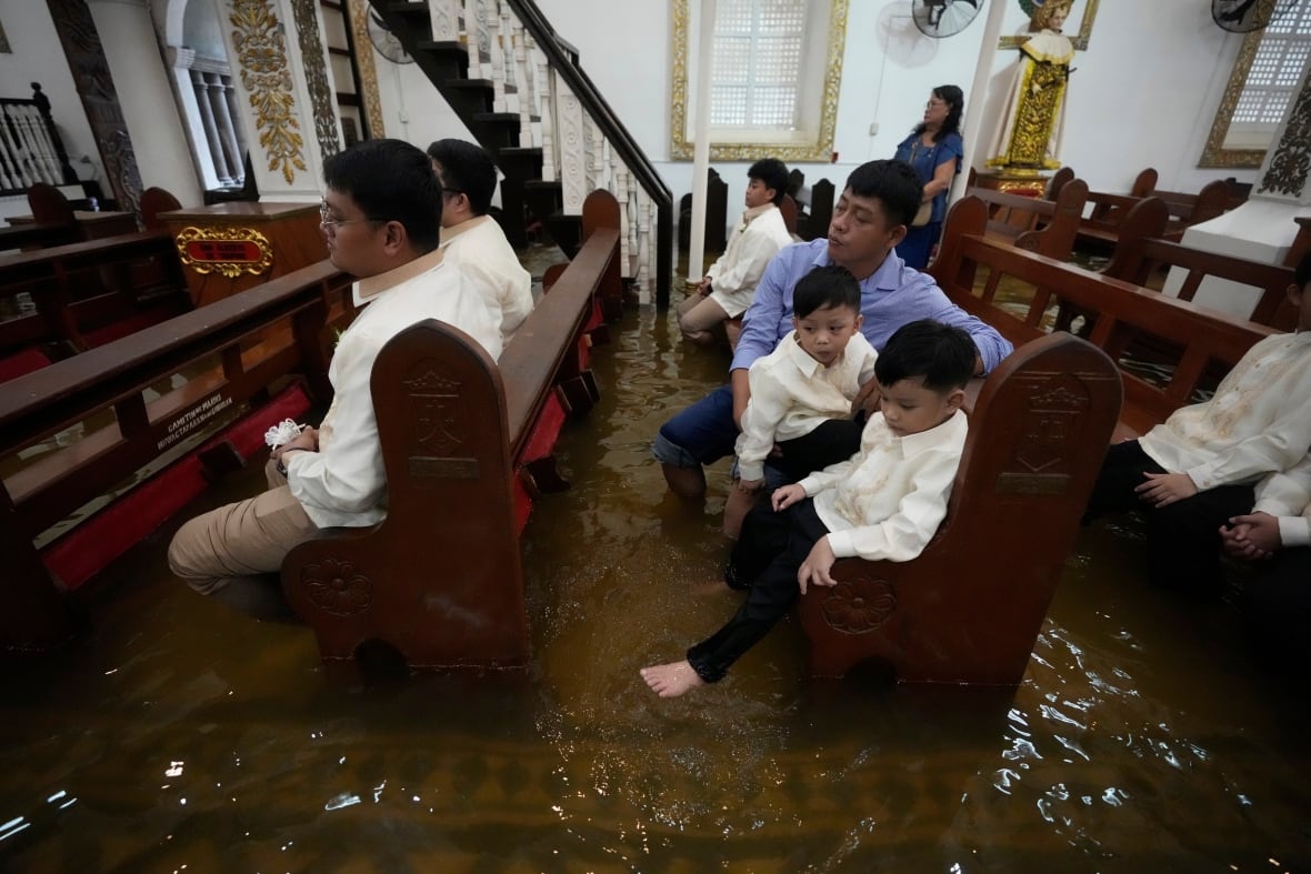 Children and adults sit on brown benches while water fills the floor in a large building.
