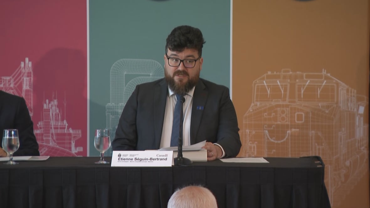 A man in a suit speaks at a table with a microphone and a nameplate reading "Étienne Séguin-Bertrand." Background panels show industrial illustrations